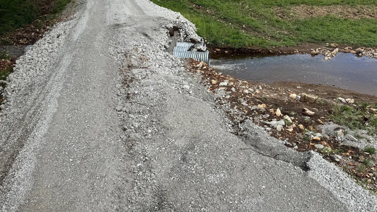 Gravel driveway over twin corrugated metal culvert pipes, its edge washing out where ditch water runs past