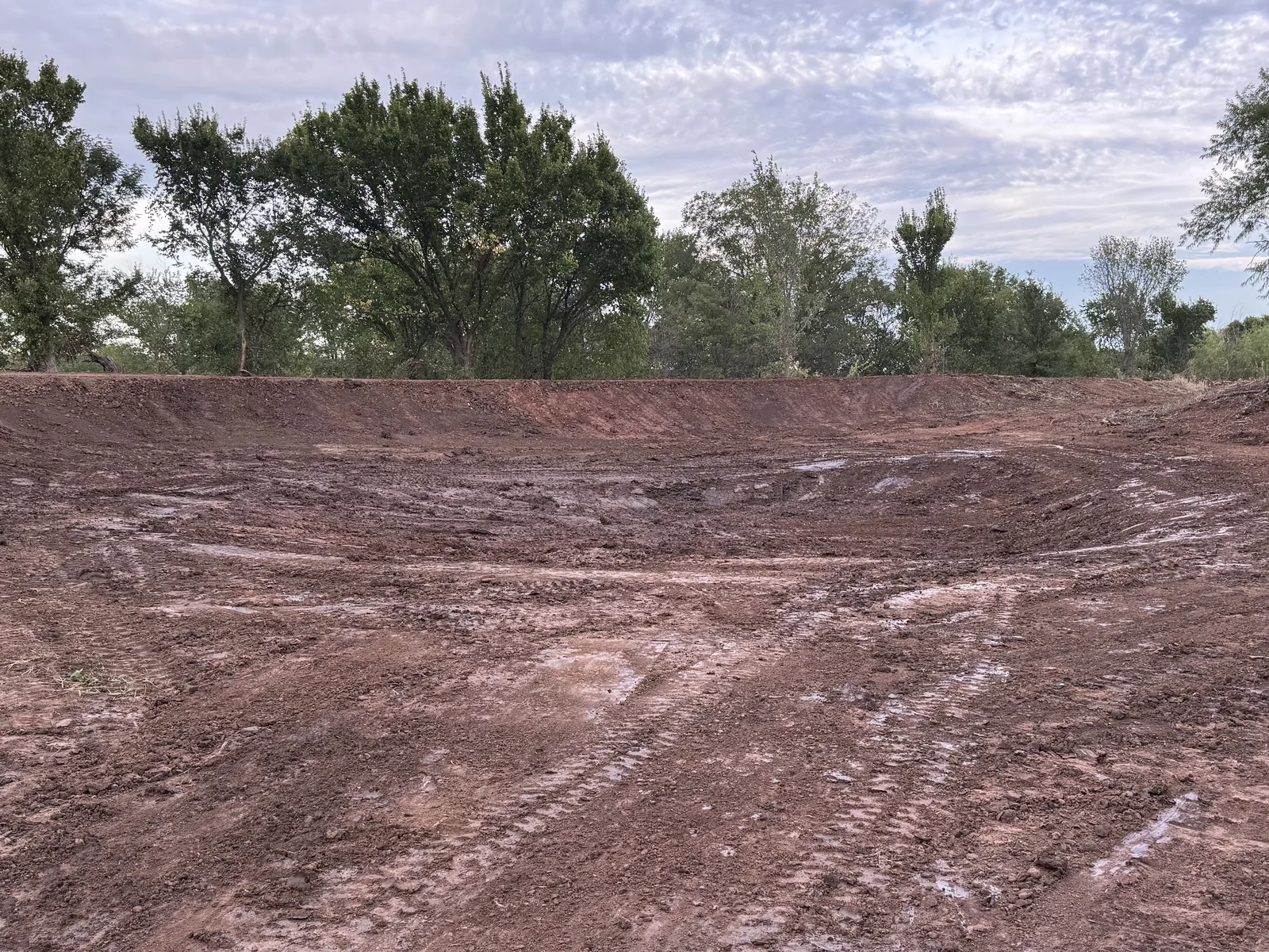 Freshly excavated pond basin cut into red clay, slopes shaped and the tree line behind