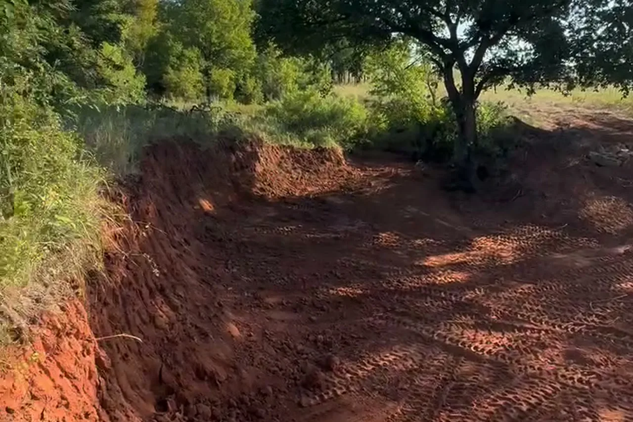 Freshly dug stock pond basin cut into red Oklahoma clay, with track loader marks across the bottom and trees on the bank